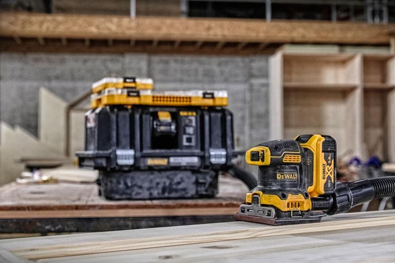 A DEWALT cordless sander (DCW200P2) is placed on a wooden surface, with a DEWALT tool storage box in the background. The