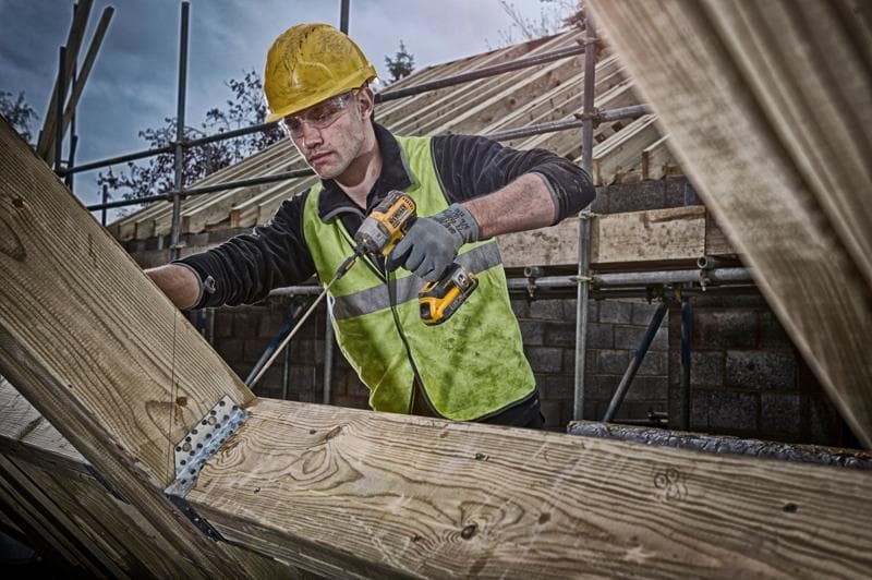 A construction worker wearing a yellow hard hat and reflective vest uses a DEWALT cordless drill to fasten metal brackets on wooden beams at a building site. His face is blurred.