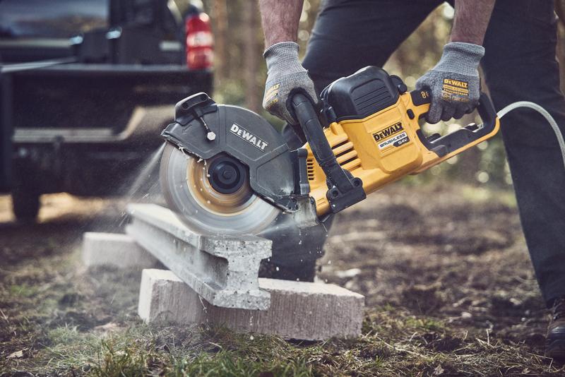 A person wearing gloves is using a DEWALT cordless power saw to cut through a concrete slab outdoors. The tool is yellow and black with visible branding, and concrete dust is seen as the saw is in action.