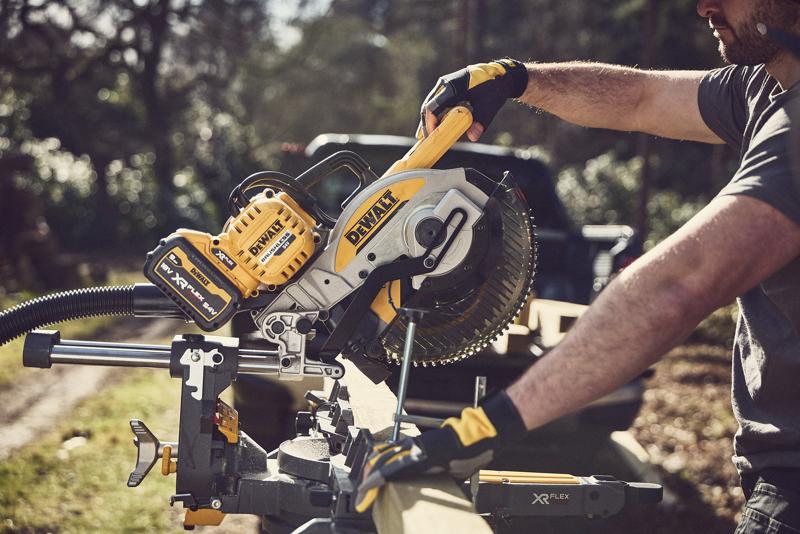 A person wearing gloves uses a DEWALT cordless sliding miter saw outdoors to cut a piece of wood. The saw features a yellow and black design and is branded with DEWALT XR FLEXVOLT.