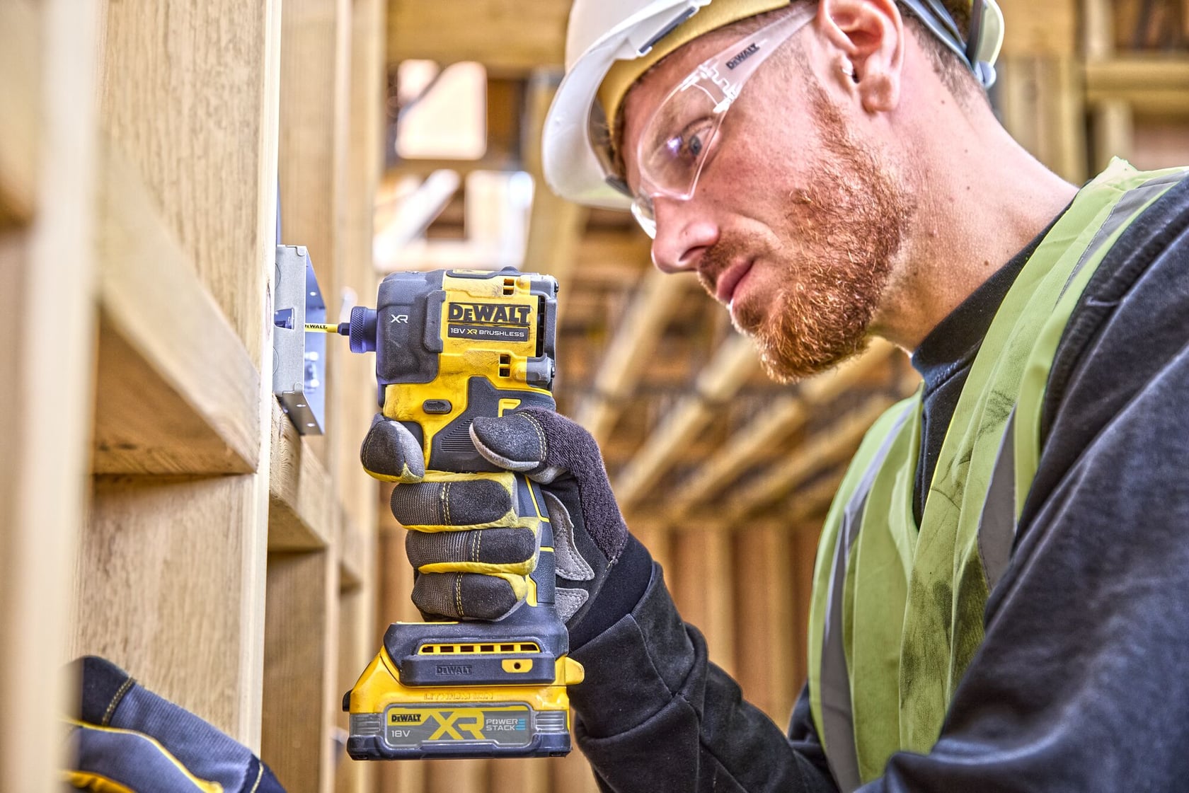 A person wearing a safety vest, gloves, and a hard hat is using a DEWALT XR cordless impact wrench (model DCF870NT) to fasten a metal bracket onto wooden framing at a construction site. The person's face is blurred.