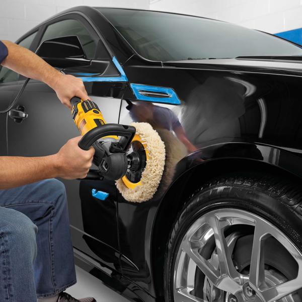 A person is using a yellow and black electric polisher with a wool pad to polish the fender of a shiny black car. Blue tape covers some parts of the car to protect them during the polishing process.