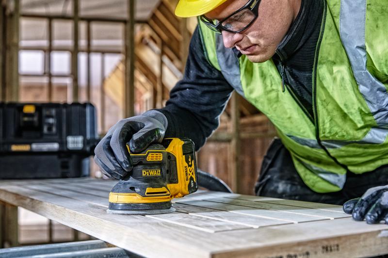 A person wearing work gloves and a high-visibility vest uses a DEWALT cordless palm sander (model DCW210) to sand a wooden board in a construction site. A black DEWALT toolbox is visible in the background.
