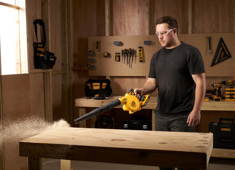 A person using a yellow DEWALT cordless blower to clean sawdust from a wooden workbench in a workshop. Various tools and tool bags are organized on the wall in the background.